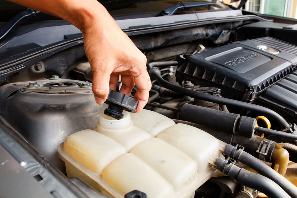 A hand opening the cap of a coolant reservoir under the hood.